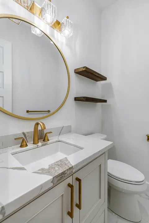 Medio baño con espejo redondo dorado, repisas flotantes de madera y llave dorada